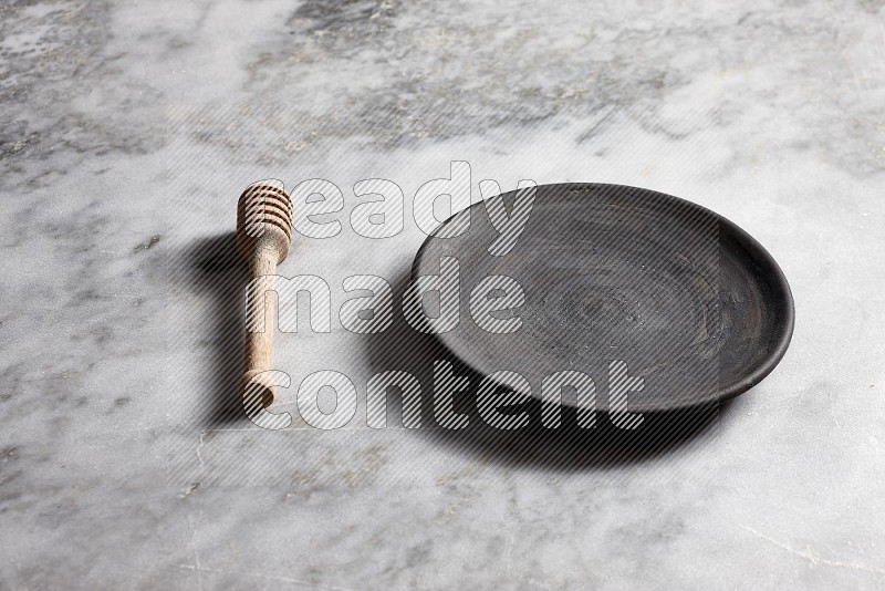 Black Pottery Plate with wooden honey handle on the side with grey marble flooring, 45 degree angle