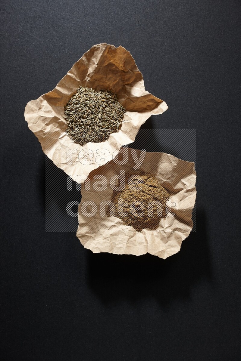 Cumin seeds and powder in 2 crumpled pieces of paper on black flooring