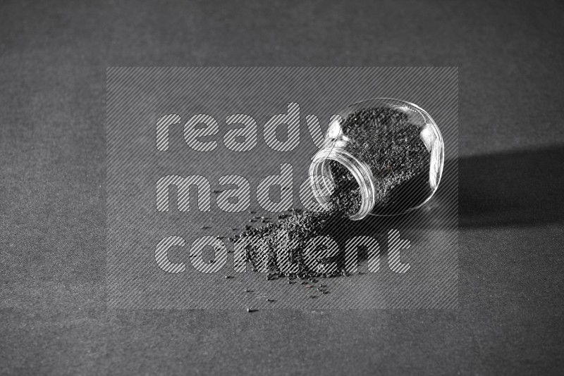 وعاء بهارات زجاجي مقلوب ممتلئ ببذور حبة البركة والبذور متناثرة بجانبه على أرضية سوداء