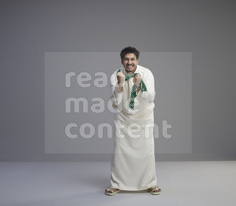 رجل سعودي يرتدي ثوب ابيض ويحمل شال اخضر على خلفية رمادي