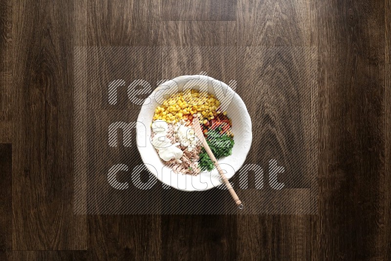 A white bowl full of tuna, colored bell pepper, sour cream, corn, parsley, black pepper powder and sauce, with wooden spoon on wooden background