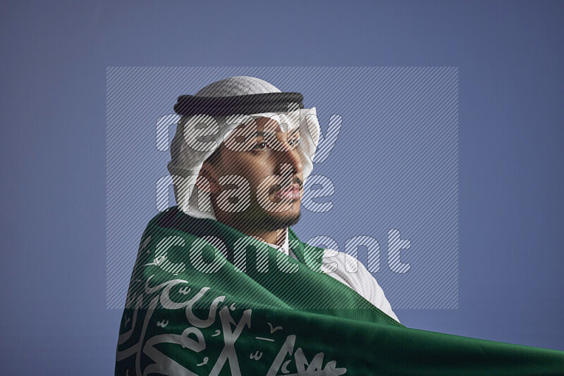 A close-up shot of Saudi man wearing thob and white shomag wrapping big Saudi flag on gray background