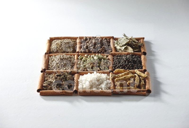 9 squares of cinnamon sticks full of cardamom in the middle surrounded by turmeric, salt, dried basil, white pepper, cumin, allspice, bay laurel leaves and black pepper on white flooring