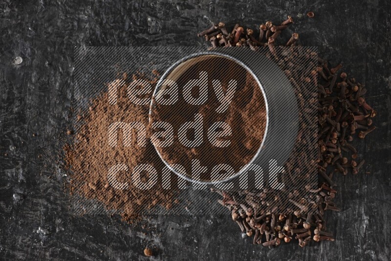 A black pottery bowl full of cloves powder spilled out of it with whole grains on a textured black flooring