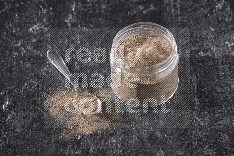 A glass jar full of black pepper powder and a metal spoon full of powder on a textured black flooring
