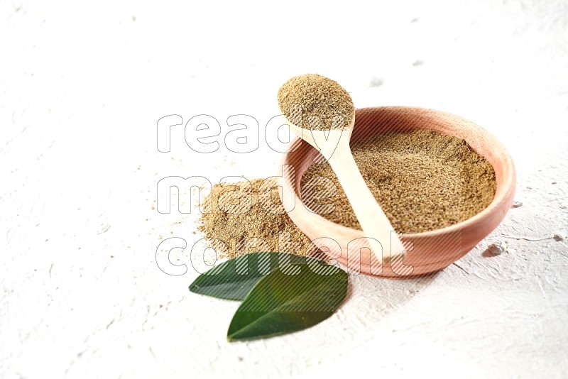 A wooden bowl and wooden spoon full of cumin powder on textured white flooring