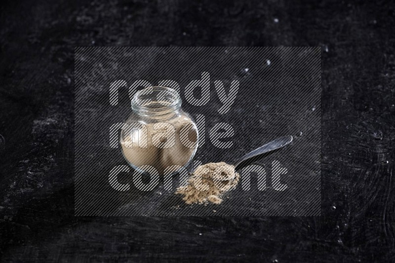 A glass spice jar full of garlic powder with metal spoon on a textured black flooring in different angles