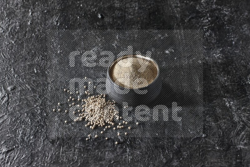 Black pottery bowl full of white pepper powder with white pepper beads on textured black flooring