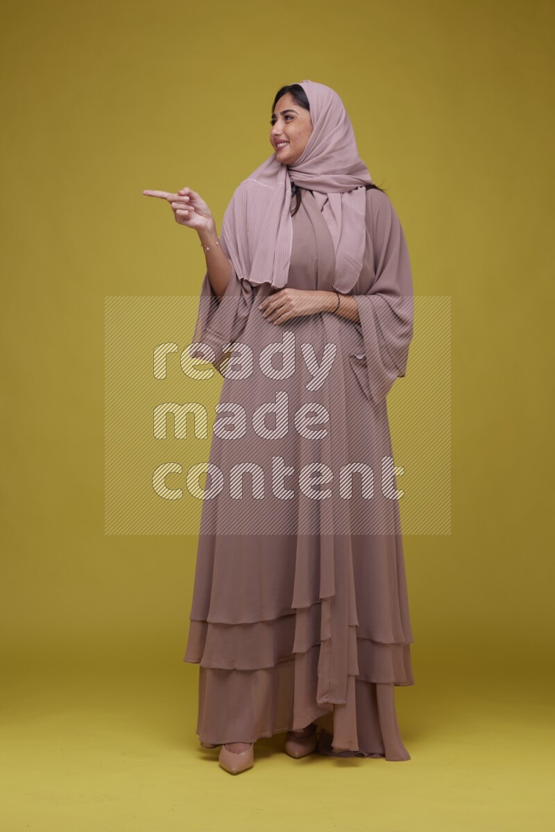 A woman Pointing on a Yellow Background wearing Brown Abaya with Hijab