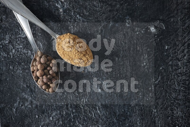2 metal spoons full of allspice powder and whole balls on a textured black flooring