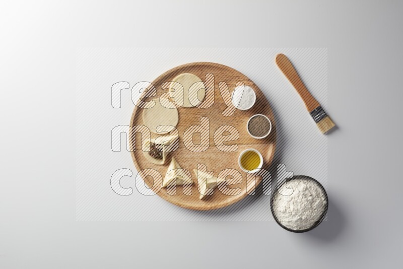 two closed sambosas and one open sambosa filled with meat while flour, salt, black pepper and oil with oil brush aside in a wooden dish on a white background
