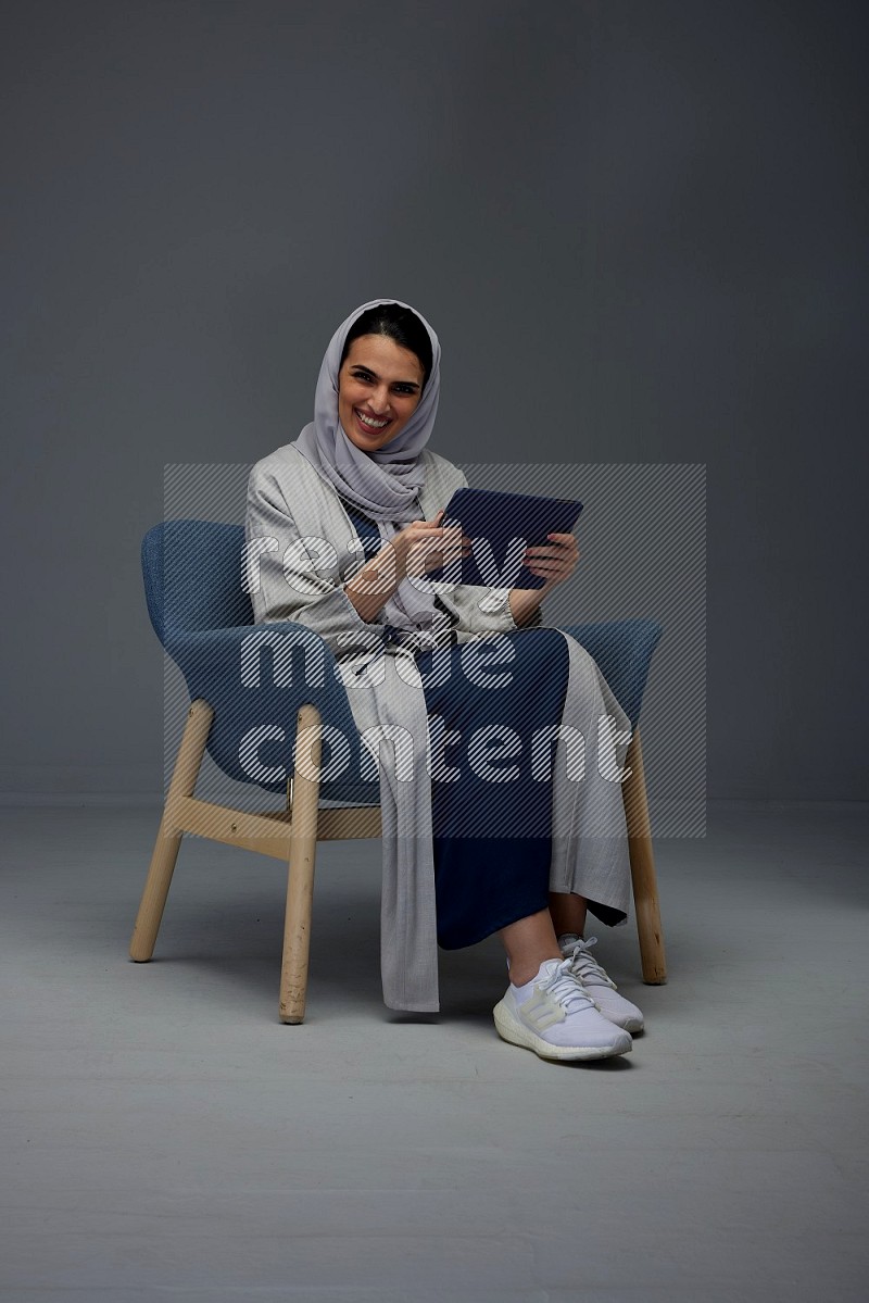 A Saudi female wearing a light gray Abaya and head scarf sitting on a dark grey chair while holding her iPad on a grey background