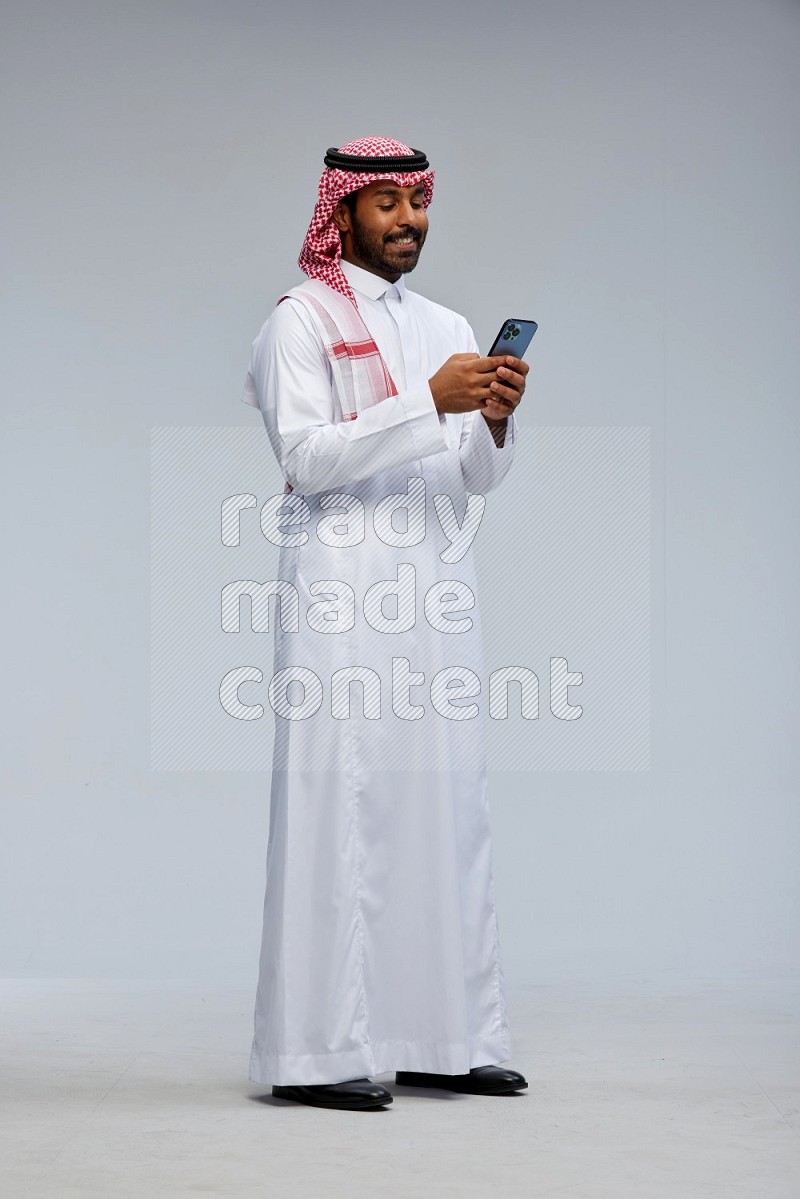 Saudi man Wearing Thob and shomag standing texting on phone on Gray background