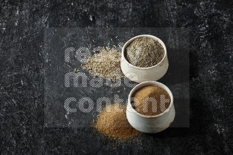 وعاءين بيج، أحدهما ممتلئ ببودرة الكمون والآخر ممتلئ بحبوب الكمون، على خلفية سوداء