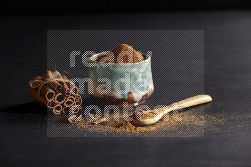 Ceramic bowl full of cinnamon powder and a wooden spoon full of powder with cinnamon sticks stacked and bounded on black background