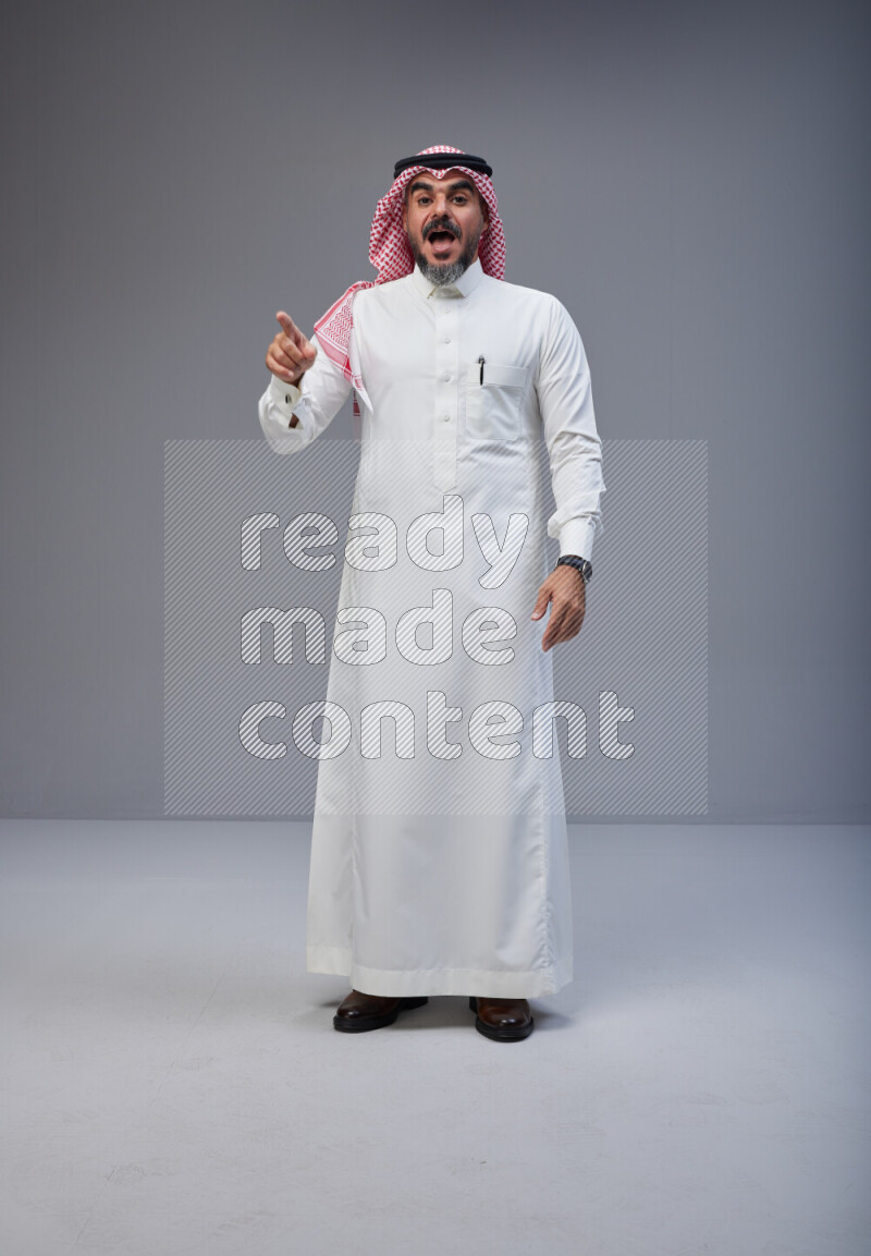 Saudi man Wearing Thob and red white Shomag standing interacting with the camera on Gray background