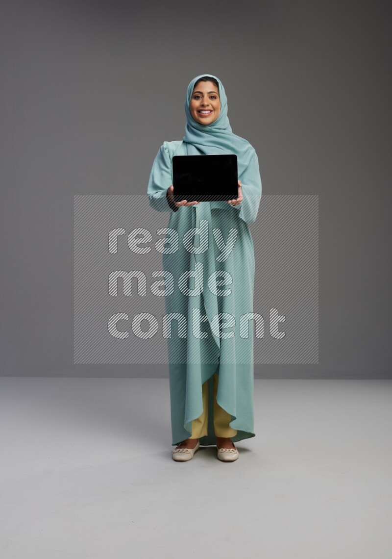 Saudi Woman wearing Abaya standing showing tablet to camera on Gray background