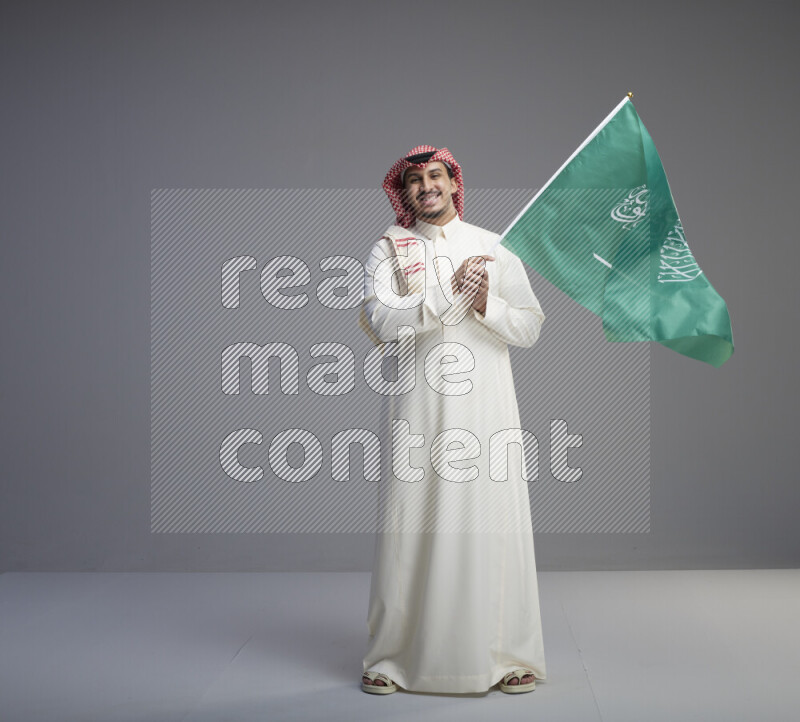 A Saudi man standing wearing thob and red shomag raising big Saudi flag on gray background