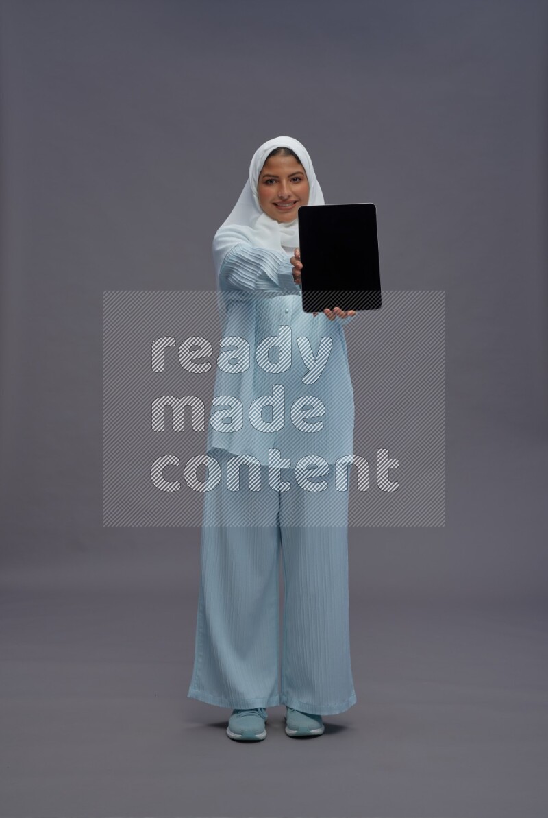 Saudi woman wearing hijab clothes standing showing tablet to camera on gray background
