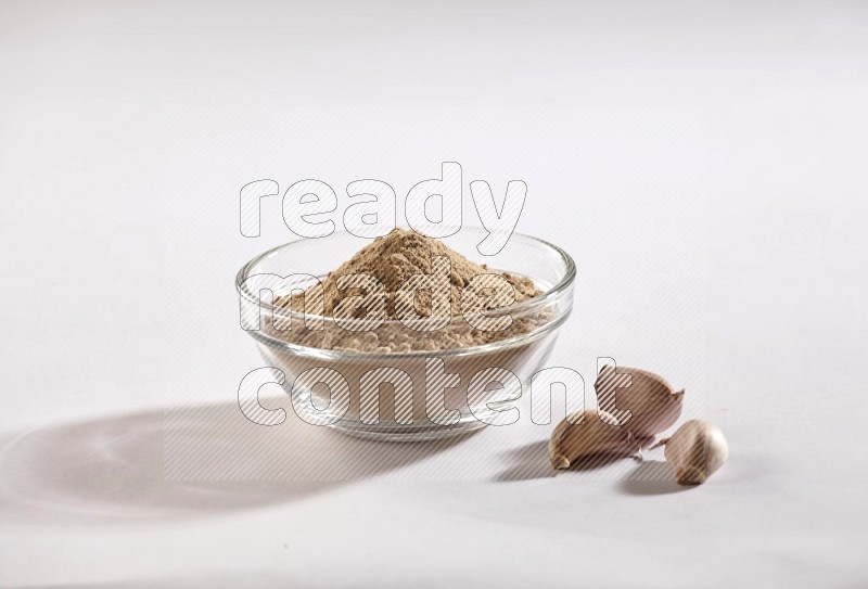 A glass bowl full of garlic powder and beside it garlic bulb and cloves on a white flooring in different angles