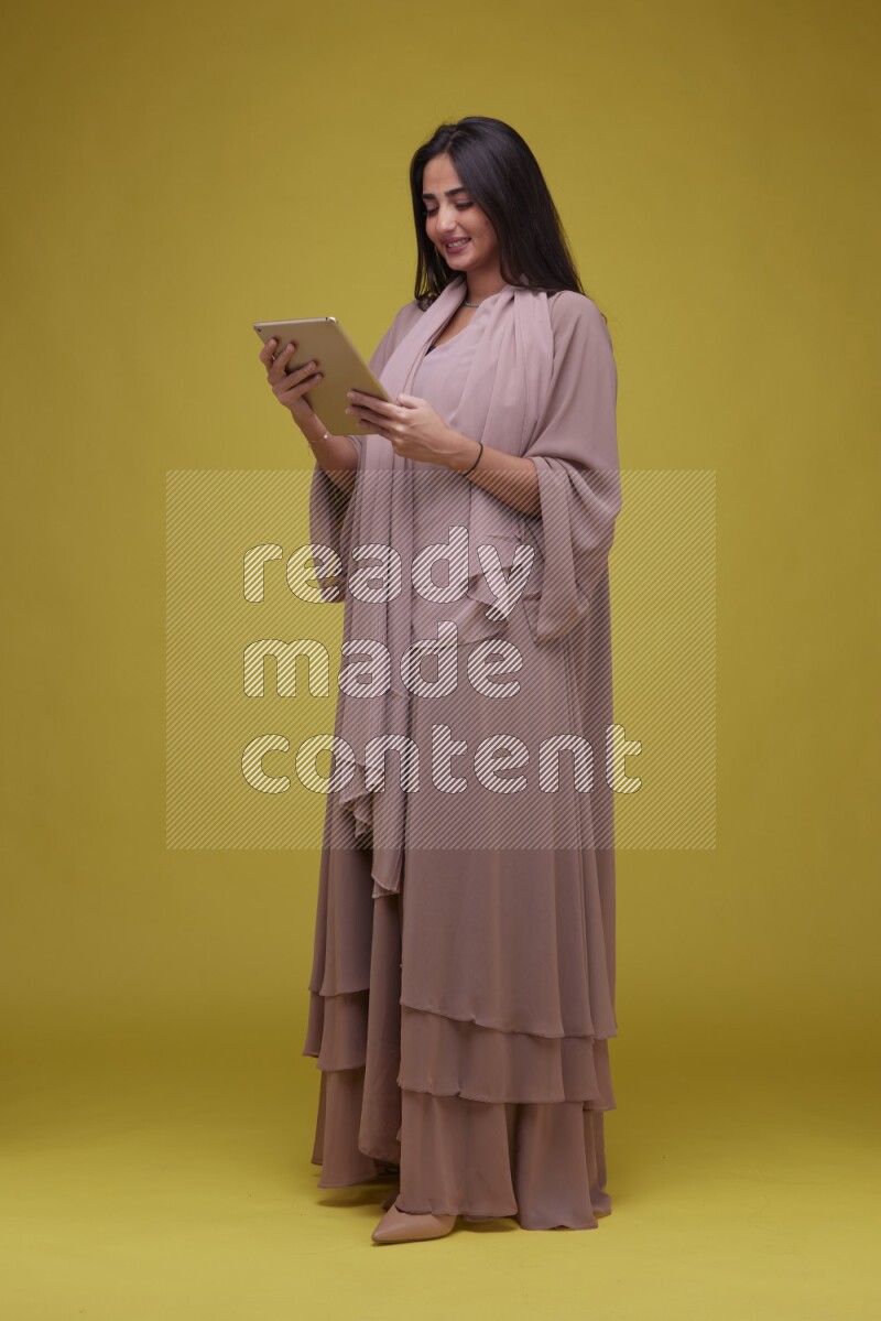 A Saudi woman Holding an iPad  on a Yellow Background wearing Brown Abaya