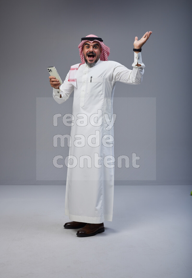 Saudi man Wearing Thob and red Shomag standing texting on phone on Gray background