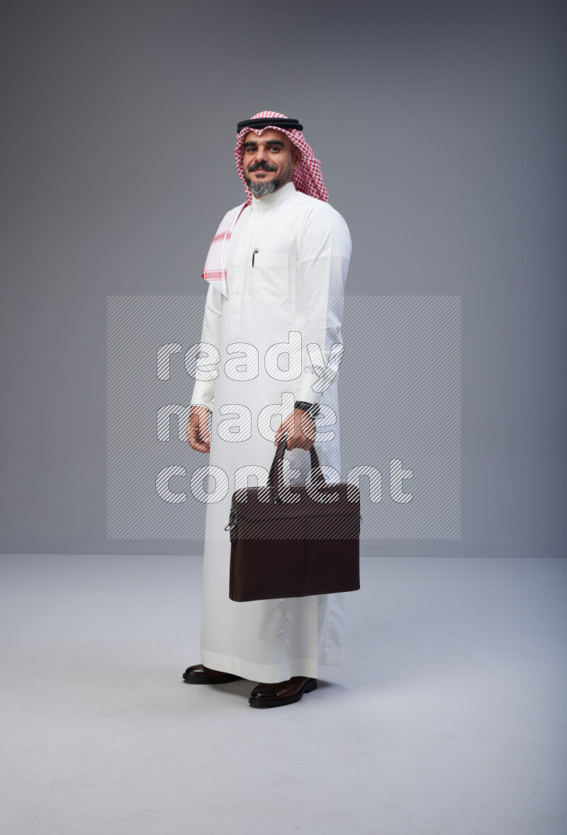 Saudi man Wearing Thob and red Shomag standing holding bag on Gray background