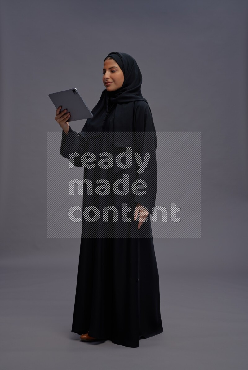 Saudi woman wearing Abaya standing working on tablet on gray background