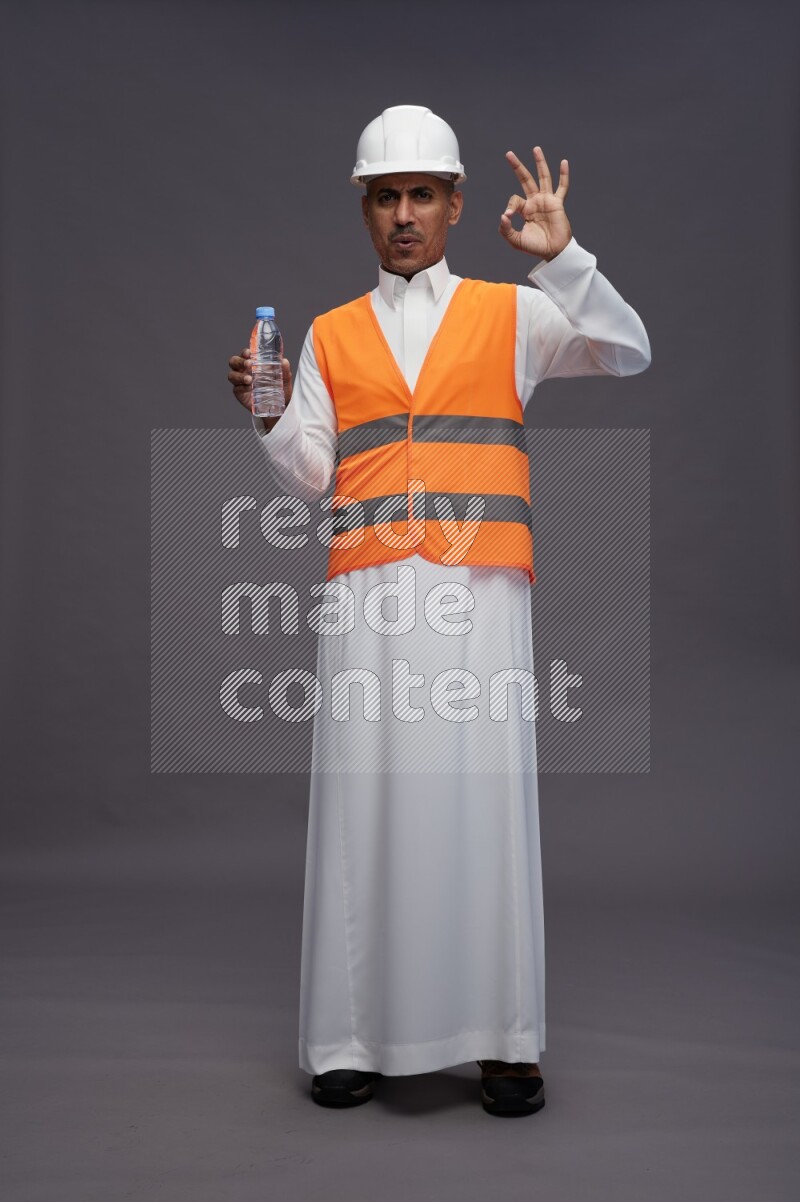 Saudi man wearing thob with engineer vest standing drinking water on gray background