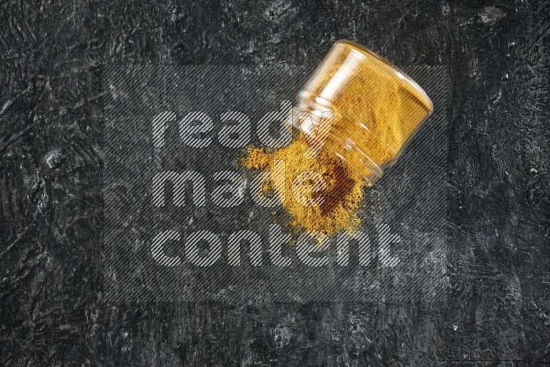 A flipped glass jar full of turmeric powder and powder spilled out of it on a textured black flooring