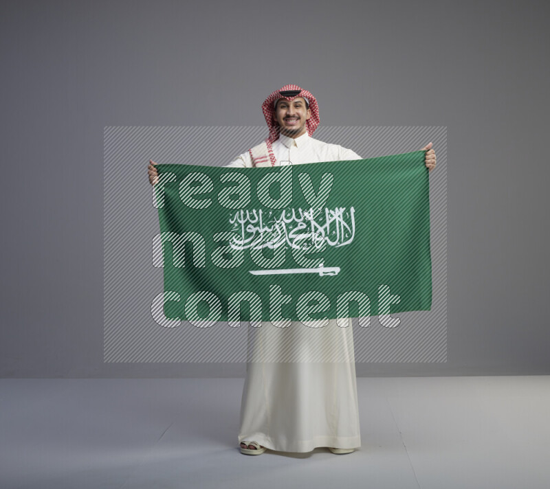 A saudi man standing wearing thob and red shomag holding big saudi flag on gray background