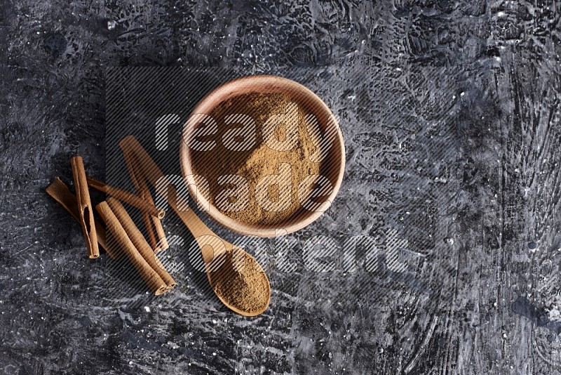 بودرة القرفة في وعاء خشبي وملعقة خشبية ممتلئة به مع أعواد القرفة على خلفية سوداء