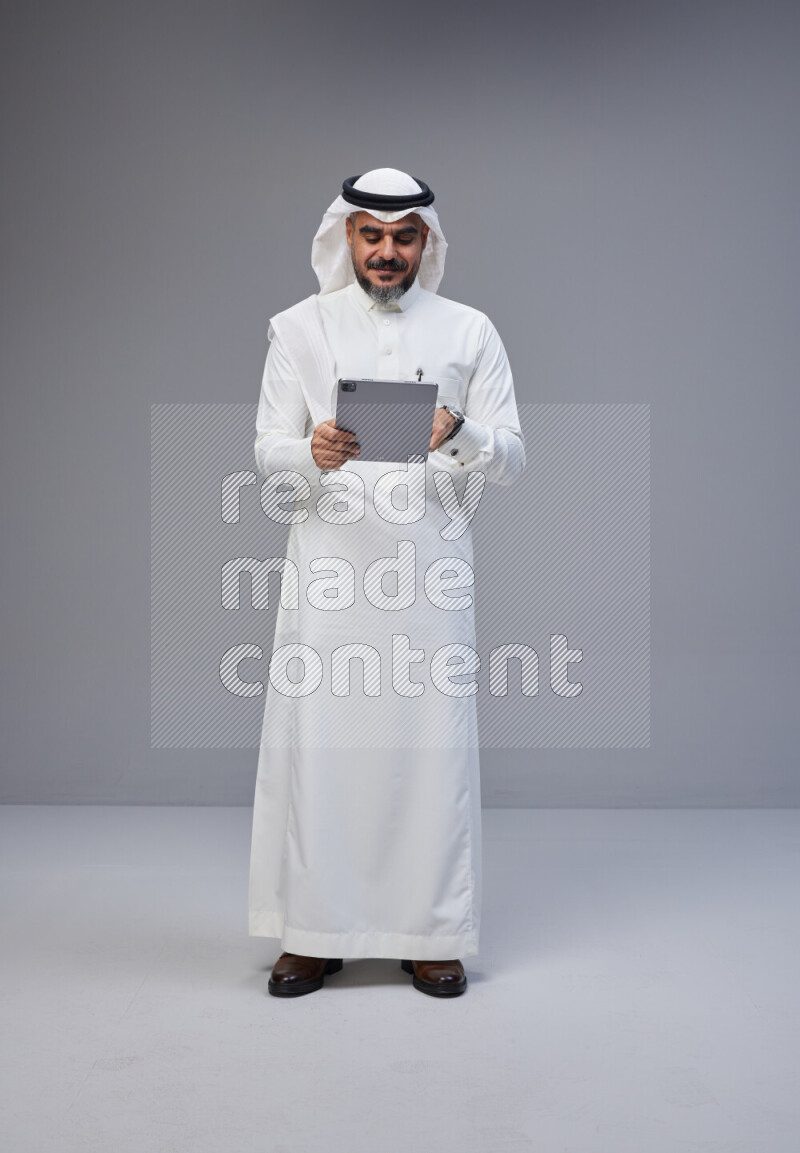 Saudi man Wearing Thob and white Shomag standing working on tablet on Gray background