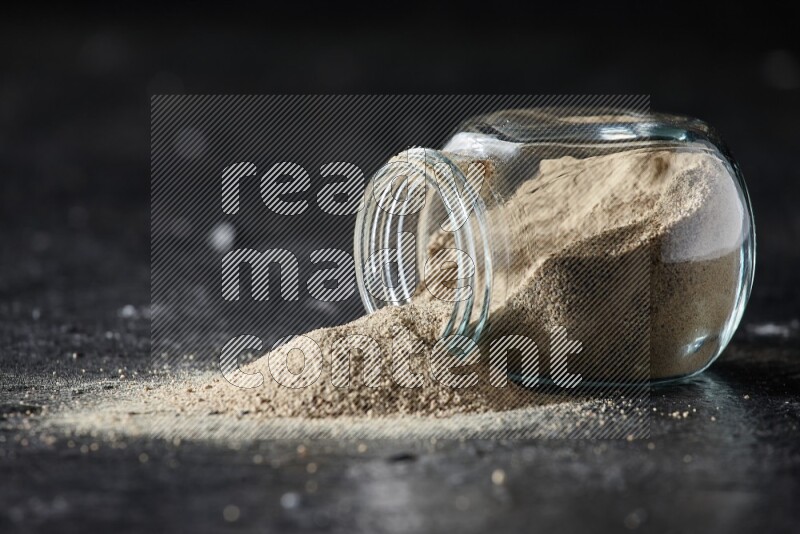 A flipped herbal glass jar full of white pepper powder with spilled powder on textured black flooring