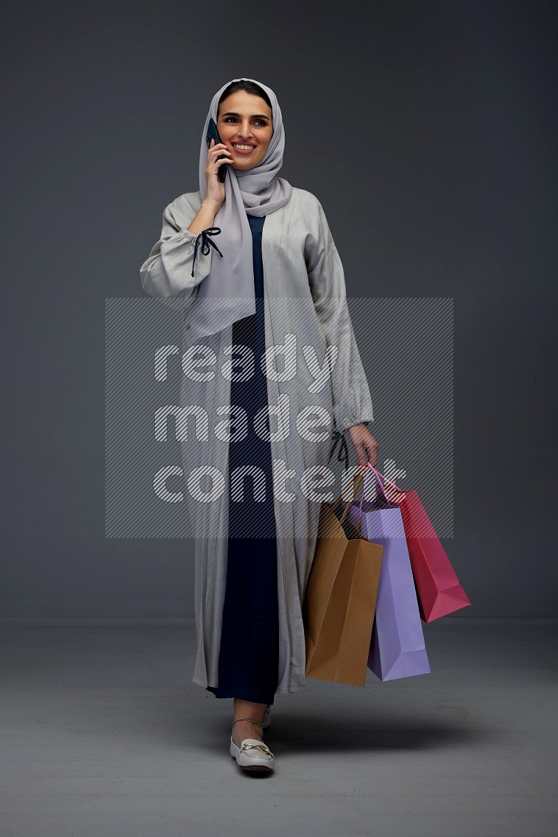 A Saudi woman wearing a light gray Abaya and head scarf standing and holding shopping bags making different poses eye level on a grey background