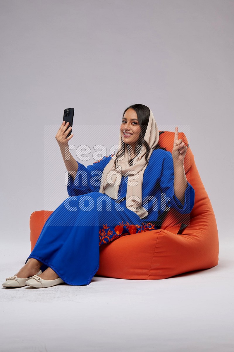 A Saudi woman wearing jalabiya sitting on an orange beanbag and taking selfie
