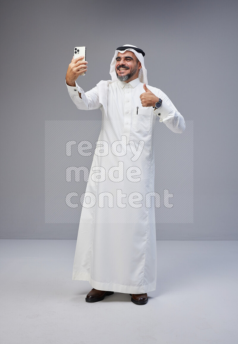 Saudi man Wearing Thob and white Shomag standing taking selfie on Gray background