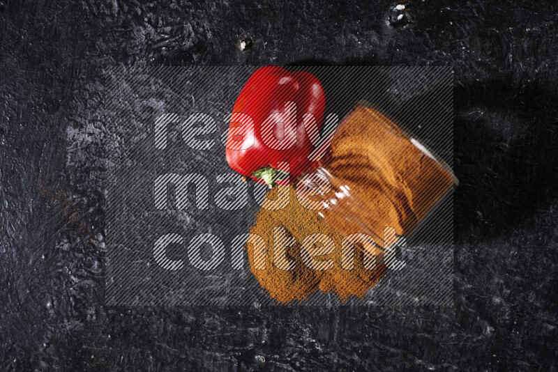 A glass jar full of ground paprika powder flipped with some spilling powder on black background