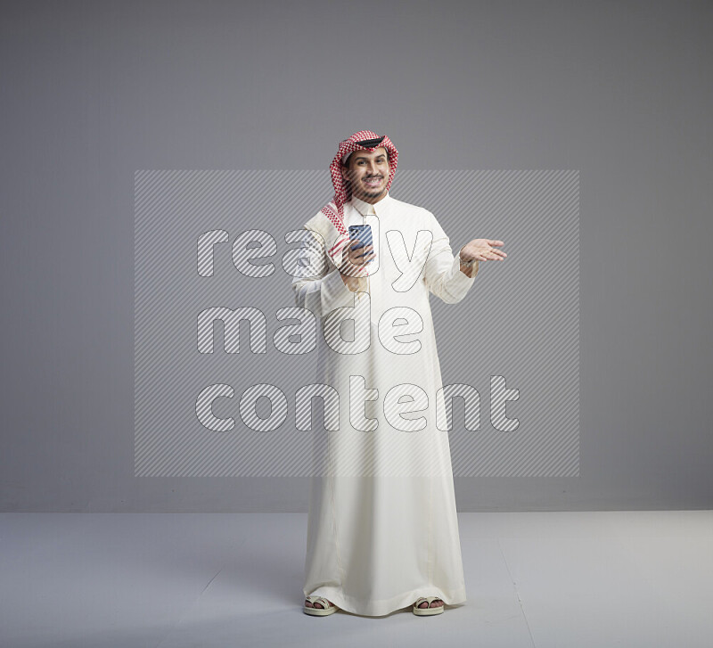 A  Saudi man standing wearing thob and red shomag texting on phone on gray background