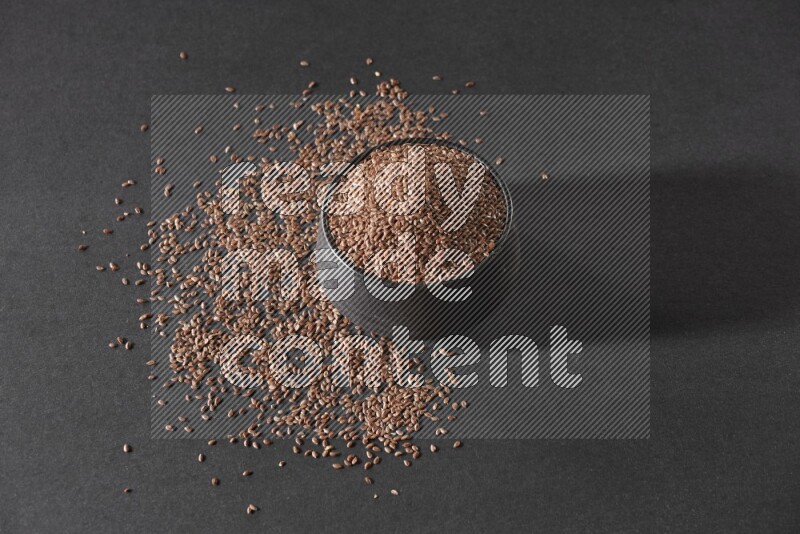 A black pottery bowl full of flaxseeds surrounded by the seeds on a black flooring
