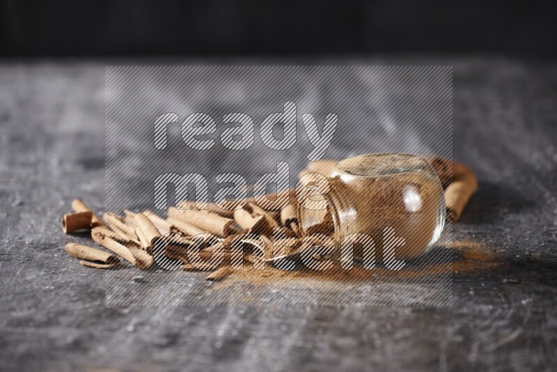 Herbal glass jar full cinnamon powder flipped and a metal spoon full of powder surrounded by cinnamon sticks on textured black background in different angles