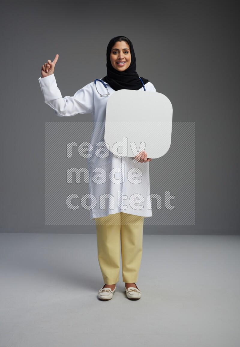 Saudi woman wearing lab coat with stethoscope standing holding social media sign on Gray background
