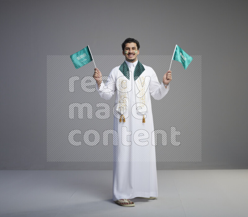 A Saudi man standing wearing thob and saudi flag scarf with face painting holding small Saudi flag on gray background