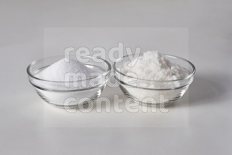 2 glass bowls one is filled with table salt and the other with coarse sea salt on white background