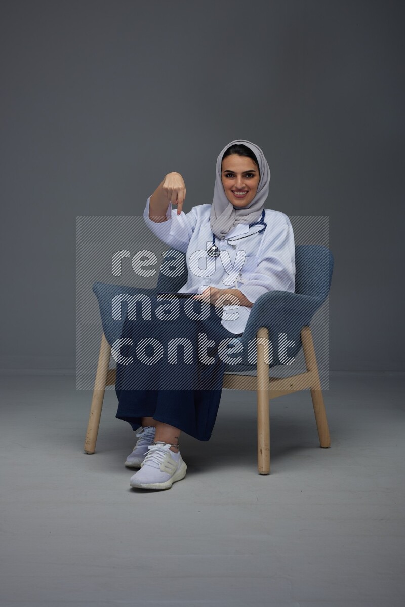A doctor wearing a light gray head scarf setting on a dark grey chair and using a phone eye level on a grey background