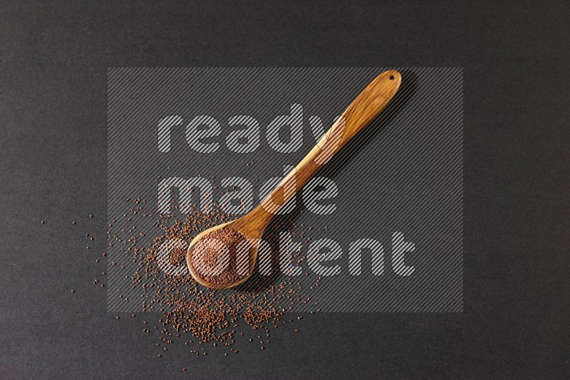 A wooden ladle full of garden cress seeds and seeds spread beside it on a black flooring