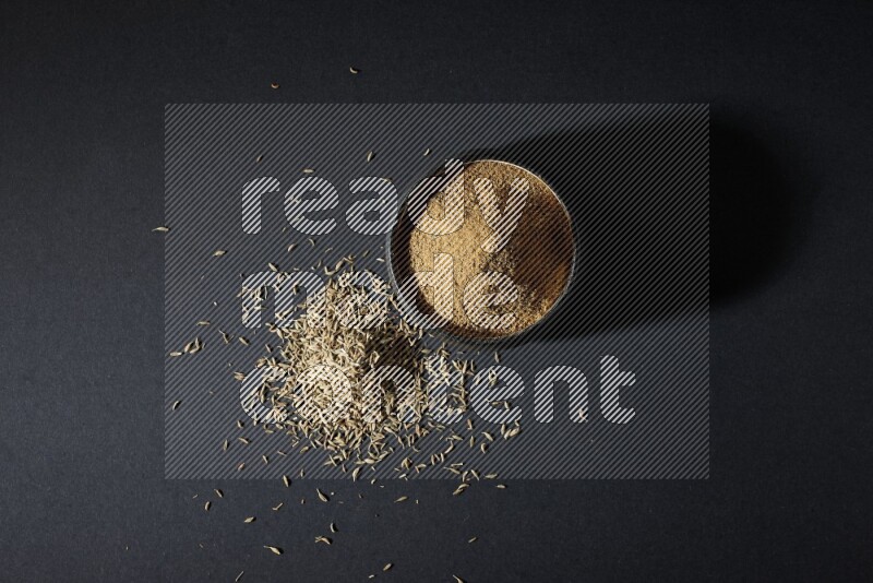 A black pottery bowl full of cumin powder and spreaded cumin seeds on a black flooring