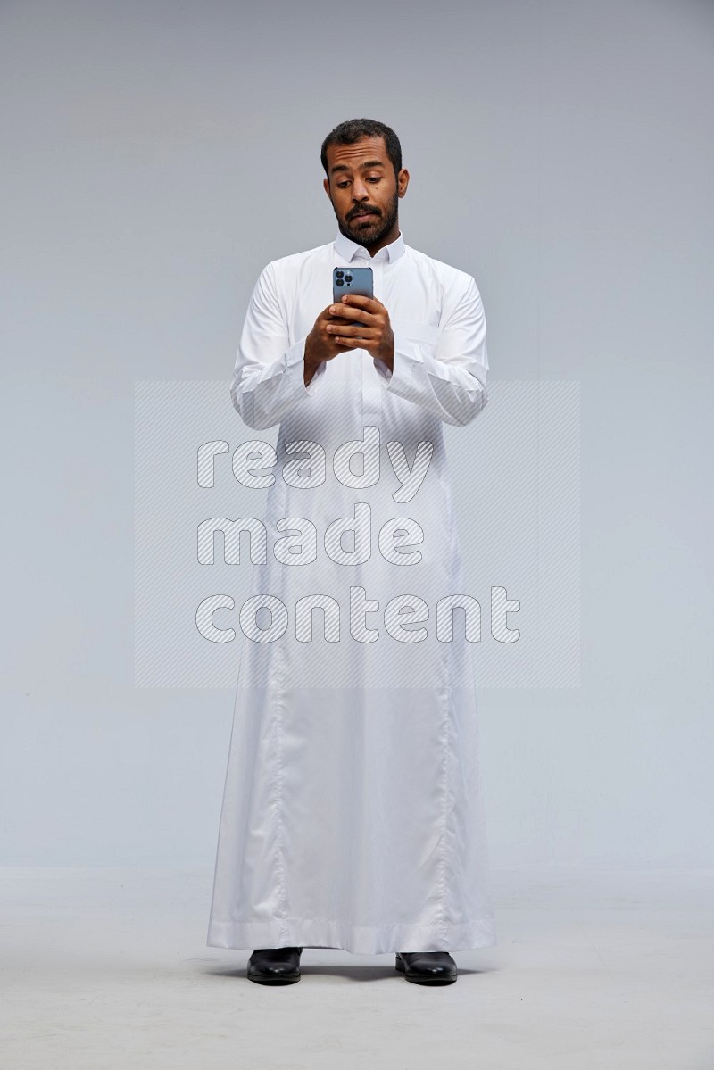 Saudi man wearing Thob standing texting on phone on Gray background