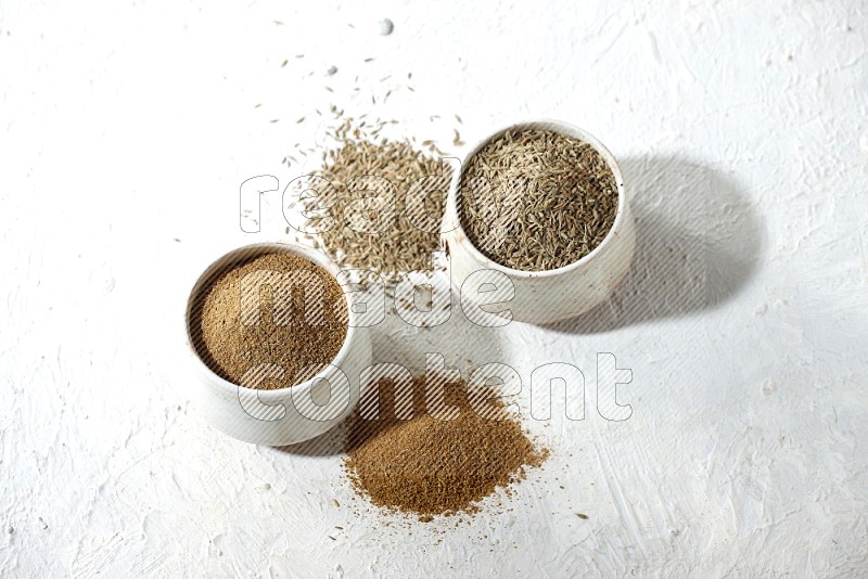 2 beige bowls full of cumin seeds and powder with spilled powder and seeds on textured white flooring