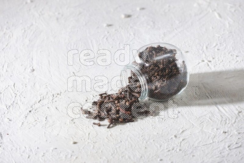 A flipped glass spice jar full of cloves and cloves came out of it on textured white flooring